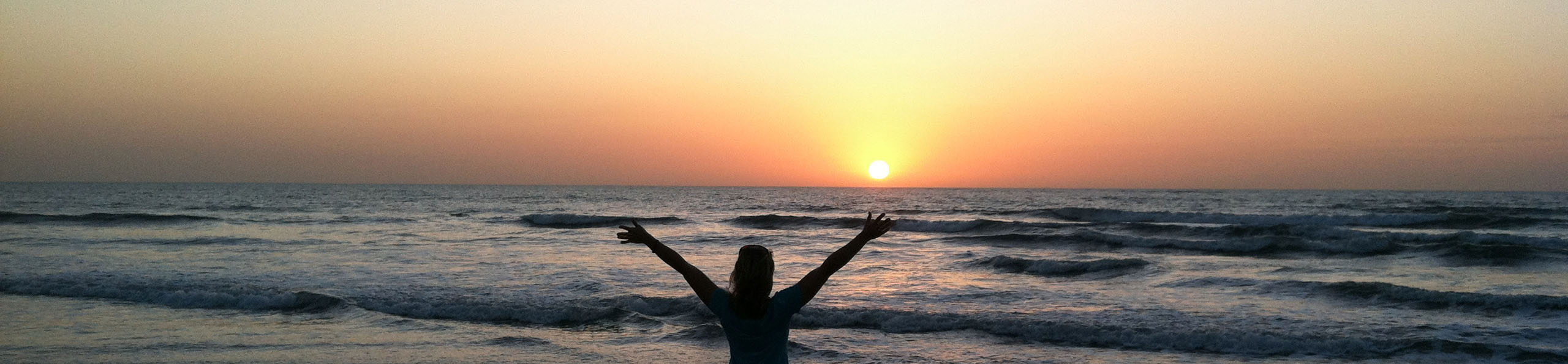 A woman with raised arms at the ocean at sunset.